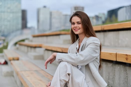 Confident woman in light business suit smiling and sitting outdoors on urban stairs with modern city buildings in background under a cloudy sky, casual business style