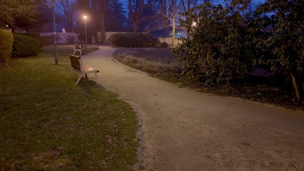 A tranquil park pathway illuminated by streetlights during twilight hours, surrounded by lush greenery.