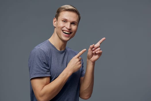 Cheerful young man with short blond hair and bright smile wearing a casual blue t-shirt, gesturing with his hands and looking joyful. The gray studio backdrop highlights his happy expression, suitable for lifestyle and happiness themes.