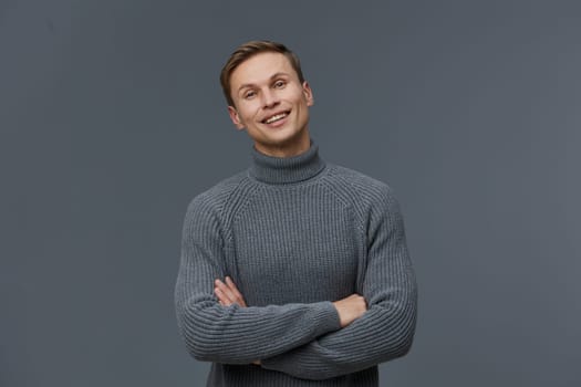 Happy young man wearing a cozy gray turtleneck sweater posing with arms crossed and smiling warmly at the camera, standing in front of a neutral gray background. Concept: confident, casual style, fashion, portrait photography, male.