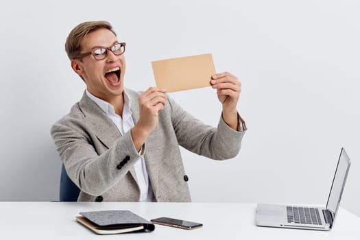 Cheerful man wearing glasses and a grey sweater holding a blank card, sitting at a modern workspace with a laptop, notebook, and calculator on a plain white background. Expressing joy, achievement, office environment, celebration, digital age, positive mood.