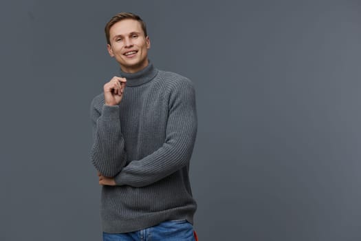 smiling young caucasian man dressed in gray knitted turtleneck sweater and blue jeans stand isolated on gray background hold hand near face happy positive emotion casual style concept digital nomad lifestyle and remote work