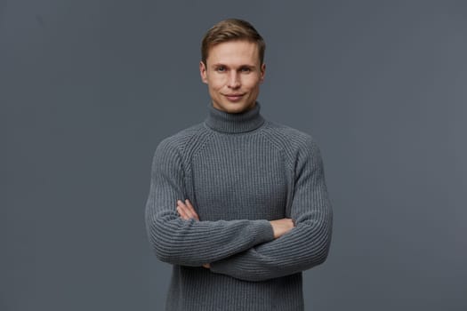 smiling man wearing gray knitted turtleneck sweater standing with arms crossed and looking at camera on dark gray background. Portrait of confident young caucasian man embodying digital nomad lifestyle, remote work, and communication technology themes. Concept: digital nomad remote work