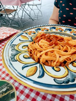 A vibrant plate of fresh pasta topped with rich tomato sauce sits on a checked tablecloth at an outdoor cafe in Italy. Surrounding tables and decor create a charming atmosphere.