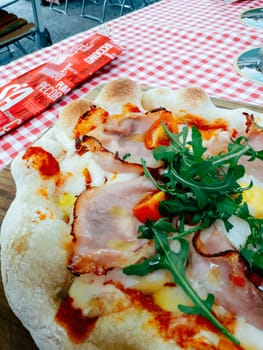 A freshly baked pizza sits on a wooden board, topped with ham, fresh tomatoes, and arugula. The vibrant red and white checkered tablecloth adds charm to the sunny outdoor market atmosphere.