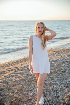 Beautiful blonde woman in summer dress walking on the beach by the sea