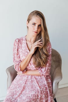 Blonde woman in summer pink dress sitting on chair in white room