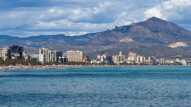 A stunning coastline features modern architecture alongside majestic mountains under a vibrant blue sky