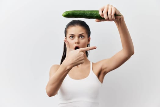 shocked woman pointing finger at cucumber while holding it above her head, displaying an expression of surprise, set against a light neutral background, capturing humor and creativity