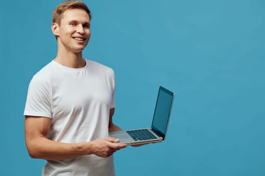 Confident young man enjoying using a laptop computer, standing against a solid blue wall. Casual white t-shirt, cheerful demeanor, digital technology, workspace, lifestyle, communication, modern environment concept.