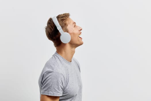 Happy young man with short brown hair and bright expression, wearing wireless headphones and a simple gray t-shirt, standing against a plain light background, conveying positivity, music enjoyment, and youthful energy. Concept: joy of music listening