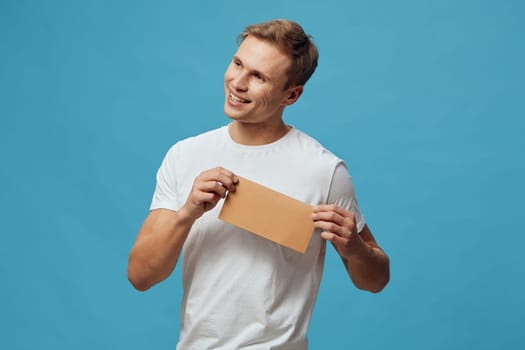 Smiling young caucasian man wearing white t-shirt holding kraft envelope and looking aside with joyful expression isolated on blue background. Bright casual studio portrait symbolizing mental health and gadgets awareness concept.