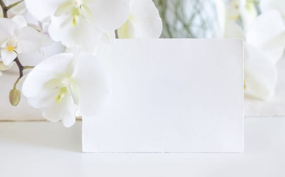 Blank card near white orchid flowers on table close up, mockup. Pastel romantic scene with horizontal card for exotic wedding, Valentines or Mothers day stationery and tropical party design