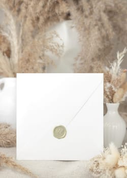 Sealed envelope against dry beige pampas grass bouquets on linen tablecloth closeup, mockup. Boho scene with blank square envelope, Bohemian or close to nature wedding stationery. Minimal composition