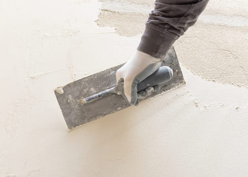 Craftsman uses a float trowel and cement to plaster a house wall. Young men with glasses in gray work clothing during construction or renovation process