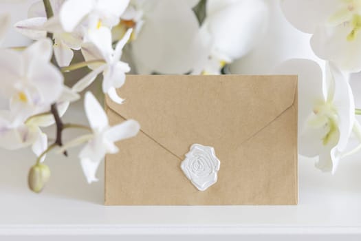 Sealed envelope near white orchid flowers on table close up, mockup. Pastel romantic scene with horizontal card for exotic wedding, Valentines or Mothers day stationery and tropical party design