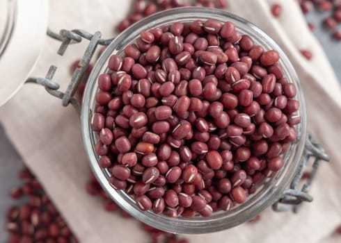Opened glass jar full of dried red azuki beans on folded napkin on gray table top view. Vegetarian protein source from legumes. Traditional East Asian cousin ingredient