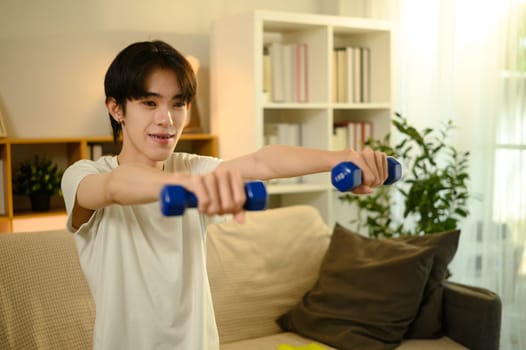 Smiling teenage man lifting blue dumbbells during a home workout.