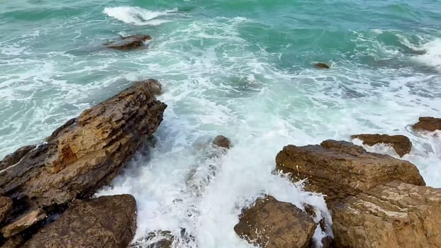 Waves gently crash and collide against the rugged, weatherworn rocks along a tranquil, turquoise coastline