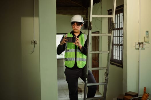 Engineer in safety vest and hard hat photographing project progress with smartphone.