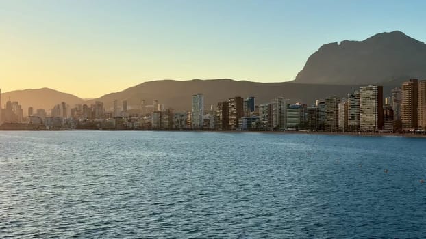 A stunning view of a coastal city skyline at sunset, with mountains in the background and serene waters.