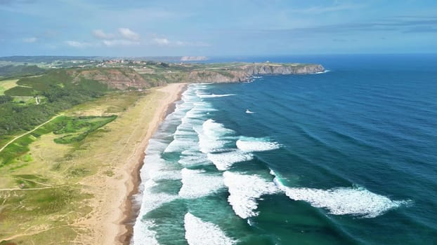 An aerial view shows a stunning coastline with crashing waves, greenery, and sandy beaches under a blue sky