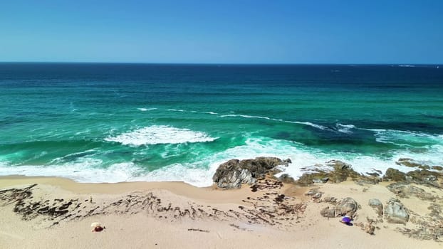A breathtaking view of turquoise waves crashing forcefully on a soft sandy beach, embraced by rugged rocky landscapes