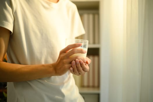 Close up of a man holding a glass of milk while standing indoors in warm natural light.