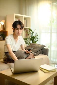 Young man relaxing on sofa while using laptop and holding smartphone in living room.