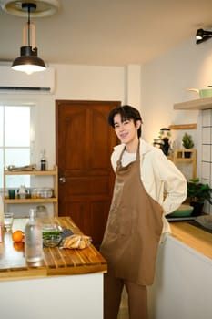 Young man wearing apron smiling in modern kitchen, ready to cook a meal at home..