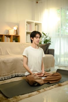 Calm male in casual attire meditating peacefully on yoga mat in living room.