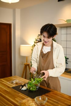 Smiling male in apron preparing a fresh green salad in a modern kitchen.