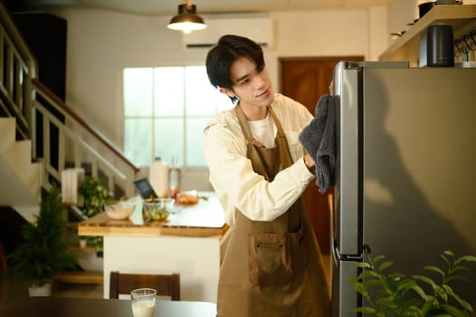 Smiling man doing household chores, cleaning the refrigerator with a cloth.