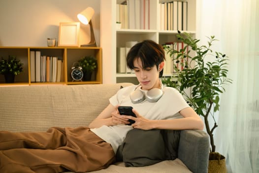 Teenage asian man lying on a sofa and using smartphone in a living room.