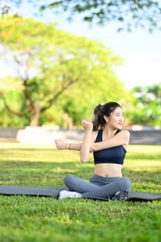 Young woman sitting on a yoga mat and stretching her arm during a workout in the park.