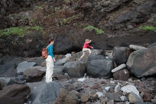 Mother watches her son jumping across rocks on a volcanic coastline