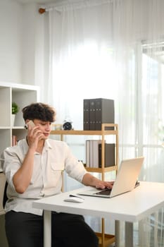 Caucasian young entrepreneur using his laptop while talking on mobile phone at workplace.