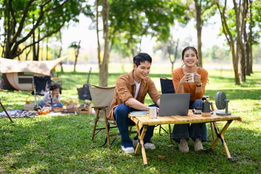 Happy couple using laptop while camping with their daughter in the background.