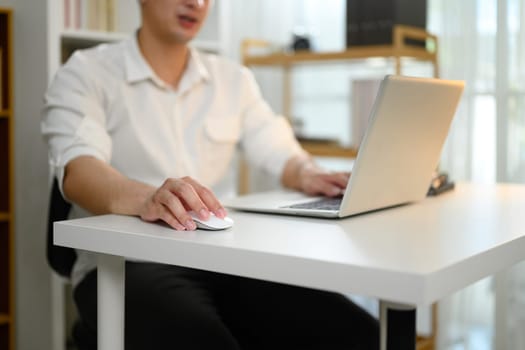 Close up of a man using wireless mouse and laptop at white desk working from home.