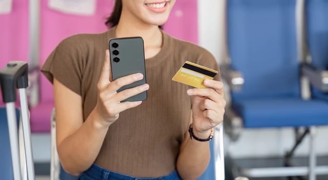 A young woman smiling while holding a smartphone and a credit card, preparing for her travel at the airport, highlighting excitement and anticipation.