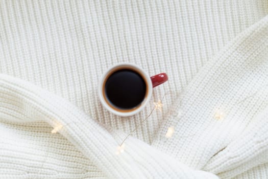 Warm coffee in a ceramic mug on a white knitted blanket with fairy lights. Cozy winter atmosphere, minimalistic holiday, Christmas, new year scene.