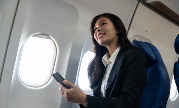A smiling businesswoman enjoying her time on the airplane while using her phone, reflecting a positive travel experience.