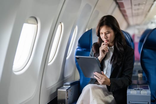 A young businesswoman looking concerned while using a tablet on an airplane, deep in thought about her plans.
