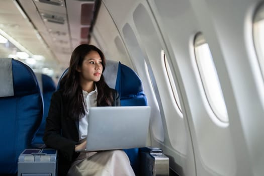A professional woman reflecting while working on a laptop, looking out of the airplane window during her business travel.