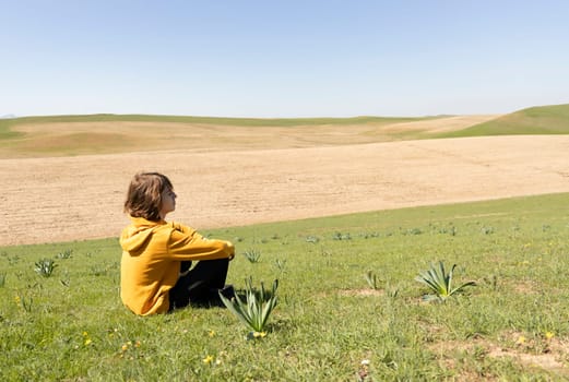Side view of a person sitting on green grass in open field, looking into the distance. Peaceful outdoor scene with copy space for text or design.