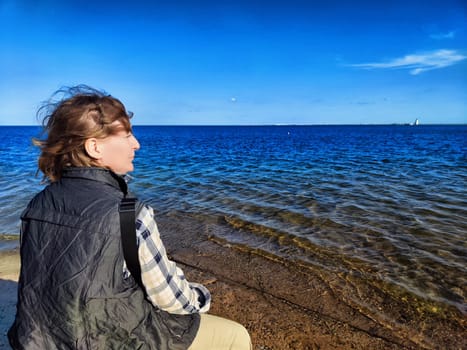 Mature woman with blonde hair relaxes by the seaside, taking in the serene ocean view on her travel excursion.