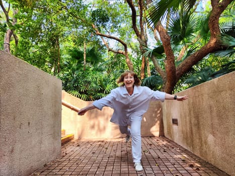 A mature blonde woman joyfully poses with arms outstretched in a vibrant tropical garden, embracing the thrill of her travel adventure.