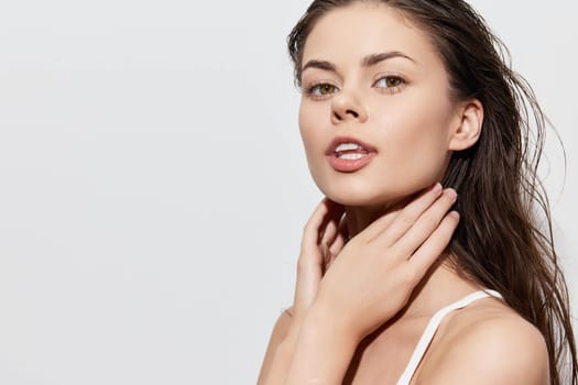 This image features a young woman with wet hair, wearing a simple white top, posing with her hand gently touching her neck The minimalist light gray background emphasizes her natural beauty and the softness of her expression, creating a serene atmosphere