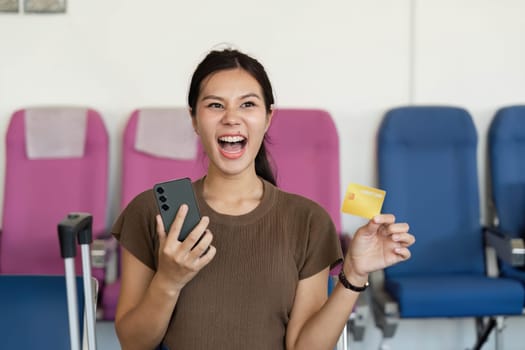 An excited young woman joyfully holding a smartphone and a credit card at the airport, ready for her upcoming travel adventure.