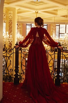 Silhouette of a model in a red dress against the background of a luxurious interior with crystal chandeliers. View from the back of an elegant young woman in a long evening dress